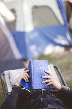 a man camping sitting outdoors reading a Bible 