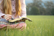 a girl sitting in a field reading a Bible 