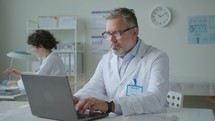 Senior Doctor Working on Laptop at Desk in Clinic
