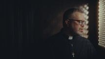 Senior Catholic priest in black robe and glasses sitting inside on confessional booth and speaking to penitent through latticed opening
