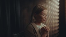 Elderly woman saying prayer with clasped hands and eyes closed in dimly lit confessional booth
