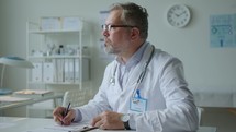 Senior Male Doctor Writing Medical Records at Workplace in Clinic
