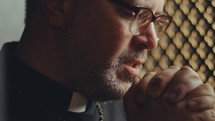 Mature priest praying deeply to God with palms together and eyes closed inside of confessional. Close-up view
