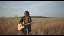 a woman standing in a field holding a guitar and singing 