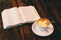 An open Bible and cup of latte' on a rustic wooden table.