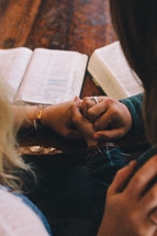 Two women holding hands and praying at a table with open Bibles.