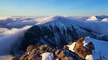 Low inversion clouds drift over snow-capped mountain peaks and hills in the valley below, showcasing the beautiful morning scenery in the national park, with rocks on the mountaintop in the foreground. PAN Camera Movement