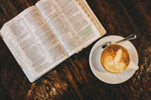 An open Bible and cup of latte' on a rustic wooden table.