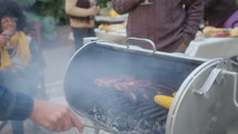 Man moving charcoal while grilling meat ribs, sausages and corn on the cob during outdoor barbecue party in the backyard. Tilt-up shot, selective focus
