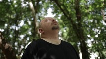 Middle aged Christian man in black shirt standing in nature, wooded tree area looking up to sky praying, meditating, worshipping.