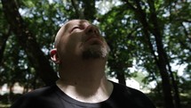 Middle aged Christian man in black shirt with beard looking up to heaven in nature while walking, hiking, meditating, praying contemplating in green, wooded area in trees with sunlight shining in cinematic, slow motion.