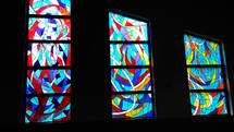 A set of three stained glass windows light a darkened corner of a church sanctuary with beautifully colored stained glass windows with red, blue, navy blue, gold, yellow, purple, violet and white stained glass colors to bring light to the dark corners of the world and add to the worship experience at local churches and chapels. 