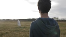 A young man, teenage boy standing in a field at sunset with sunlight shining sees Jesus Christ or angel or spiritual figure in white robe.