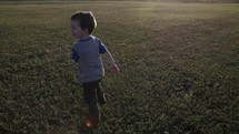 A young, cute happy boy in a blue shirt running in sunrise or sunset sunlight in green grass in cinematic slow motion.