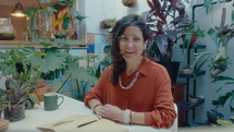 Smiling woman sitting at table in indoor garden at home, looking at camera and discussing green houseplants during web call. POV medium shot
