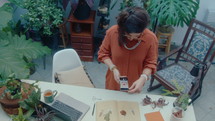 Woman standing at desk in indoor home garden and photographing handmade herbarium book with mobile phone while making content for DIY blog on social media. High angle shot
