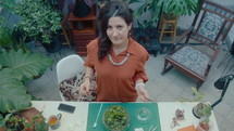 Female florist holding tweezers and scissors, standing by table with decorative terrarium on it and posing for camera in flower workshop. High angle shot, video portrait
