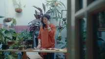 Businesswoman standing among lots of green plants in plant shop, having talk on mobile phone and using laptop while taking an order from customer
