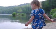 Toddler girl throws big rock into lake on bright summer morning
