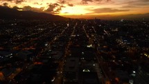 Aerial shot drone flies forward toward sun over city lit up at sunset