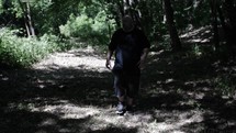 Middle aged Christian man in black shirt with beard in nature walking, hiking, meditating, praying contemplating in green, wooded area in trees with sunlight shining in cinematic, slow motion.