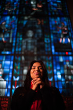 a woman with praying hands standing in front of a stained glass window 