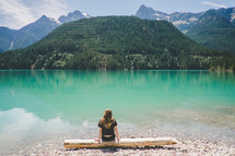 A woman sits on a log at the edge of a lake surrounded by mountains.