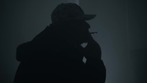 Silhouette of drug addict, man wearing a hat and hooded coat in a dark, smoky room smoking a cigarette or drugs like marijuana. 