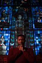 a man with praying hands in church standing in front of a stained glass window 