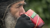 Side profile of a gray bearded man drinking out of a mug on the street.