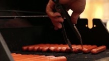 Hand with tongs turning hot dogs on a grill.