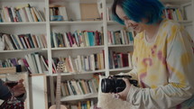 Cheerful girl with blue hair filming her boyfriend with camera when he drinking wine at home music studio, young gen Z couple having fun at leisure time
