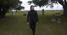 Sombre, sad young man in black suit walking in cinematic slow motion in cemetery through graveyard tombstones.