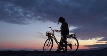 Silhouette of a Girl with magic glowing bicycle walk near the ocean at sunset sky