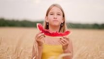 Cute girl eating watermelon standing in wheat field on sunny summer day. Child eats a delicious watermelon, summer lifestyle. Kid emotion at summer vacation.