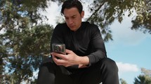 Seated Man In The Park Using Smartphone Under The Trees