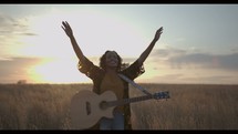 a woman standing in a field holding a guitar with her arm raised 