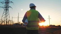 Silhouette of engineer walk on field with electricity towers. Electrical engineer with high voltage electricity pylon at sunset background. Power workers at work concept.