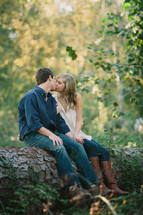 man and woman kissing while sitting on the trunk of a fallen tree