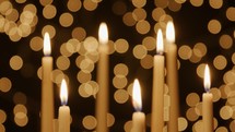 Seven lit taper candles with Christmas light bokeh in the background.