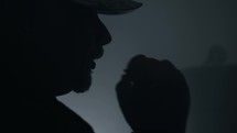 Silhouette of drug addicted man wearing a hat and hooded coat in a dark, smoky room smoking a cigarette or drugs like marijuana. 