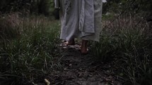 The feet of Jesus Christ wearing white robe / tunic walking in cinematic, dramatic slow motion in wooded area surrounded by trees and green grass. 