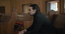 Young, emotional, anxious, and stressed man with long hair and black suit sitting in old church in worship and praying.