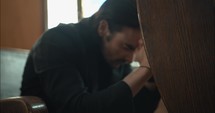 Young, emotional, anxious, and stressed man with long hair and black suit sitting in old church in worship and praying.