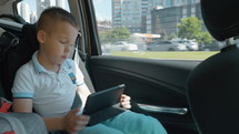 Boy traveling in the city by car. He sitting at the back seat with safety belt and watching movie on tablet computer