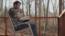 Young man sitting on front porch reading his Bible and drinking coffee