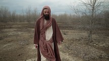 Jesus Christ or bible prophet like Noah, Moses, Elijah, Abraham or John the Baptist wearing brown robes and tunic, walking in cinematic slow motion in a dry barren wilderness.