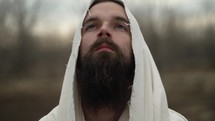 Risen Jesus Christ after the easter resurrection. Or biblical prophet or Christian man with beard dressed in white, tattered tunic with hood standing outside in front of distant trees.