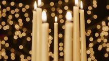 Seven lit taper candles that focuses in on one candle with Christmas light bokeh in the background.