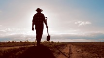 Farm agriculture concept. Farmer with a shovel in rubber boots walks across field at sunset. Silhouette man gardener walking along the road along the field. Tired man after hard work on the ground.